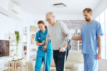 Team of nurses or social workers helping an old disabled man to walk with his crutches out of the nursing home room.