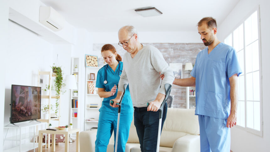 Team of nurses or social workers helping an old disabled man to walk with his crutches out of the nursing home room.