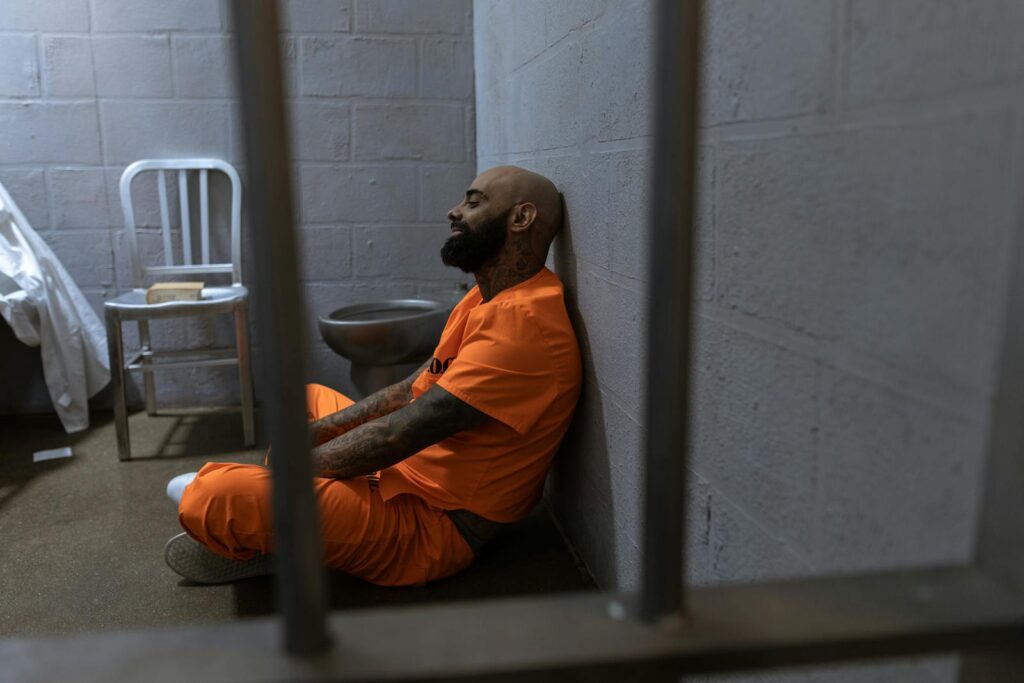 A bald man in an orange jumpsuit sits on the floor of a jail cell, leaning against a concrete wall.