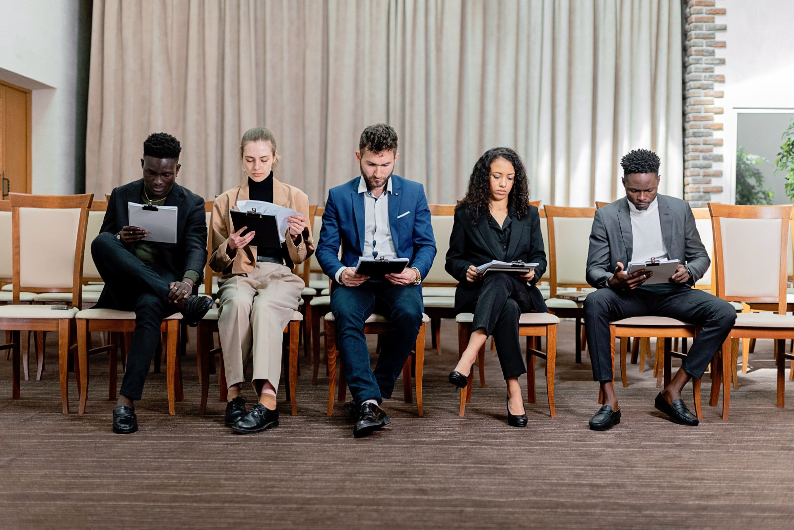 Five diverse business professionals sitting in a waiting room holding tablets and documents, representing jury duty or legal proceedings.