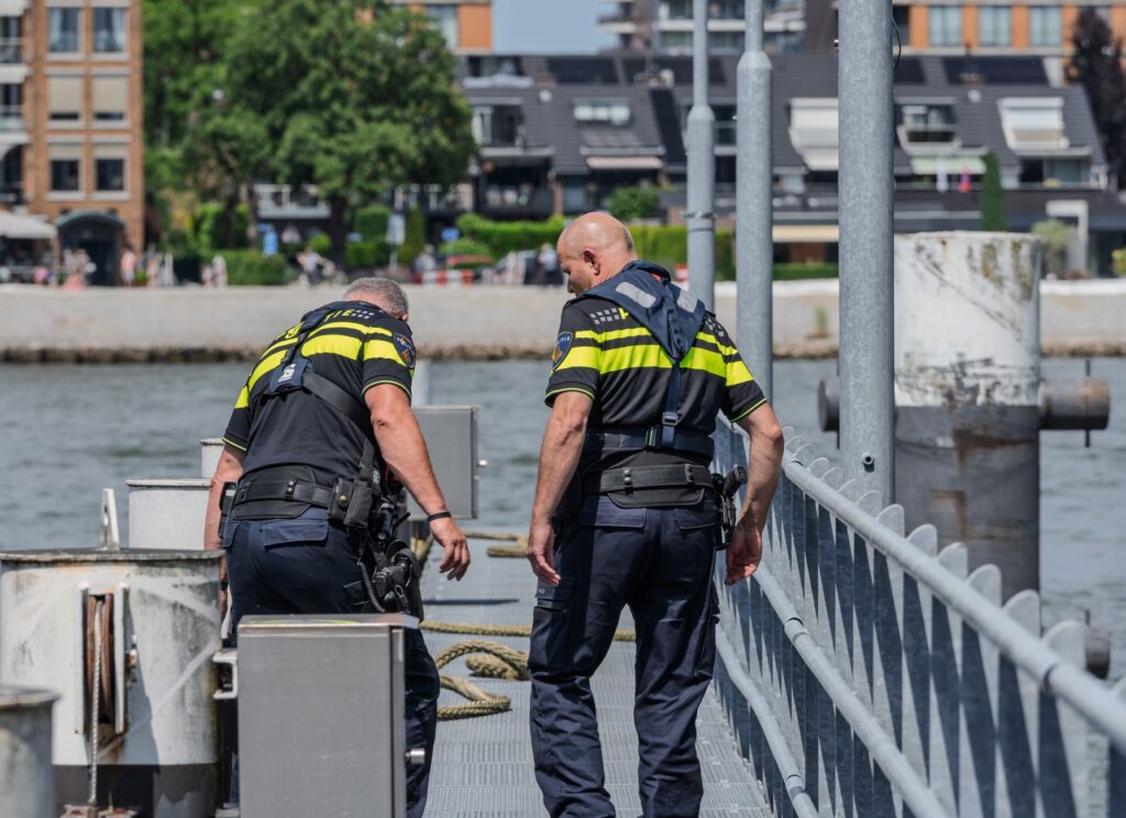 Two police officers patrol a riverside area, with buildings in the background.