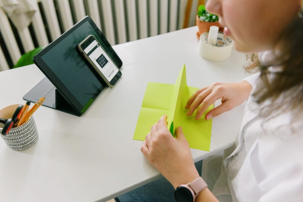 A woman folding green paper at a desk, with a tablet and phone nearby, in a cozy indoor setting.