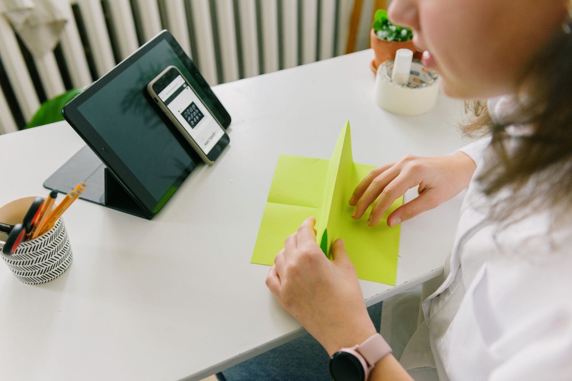 A woman folding green paper at a desk, with a tablet and phone nearby, in a cozy indoor setting.