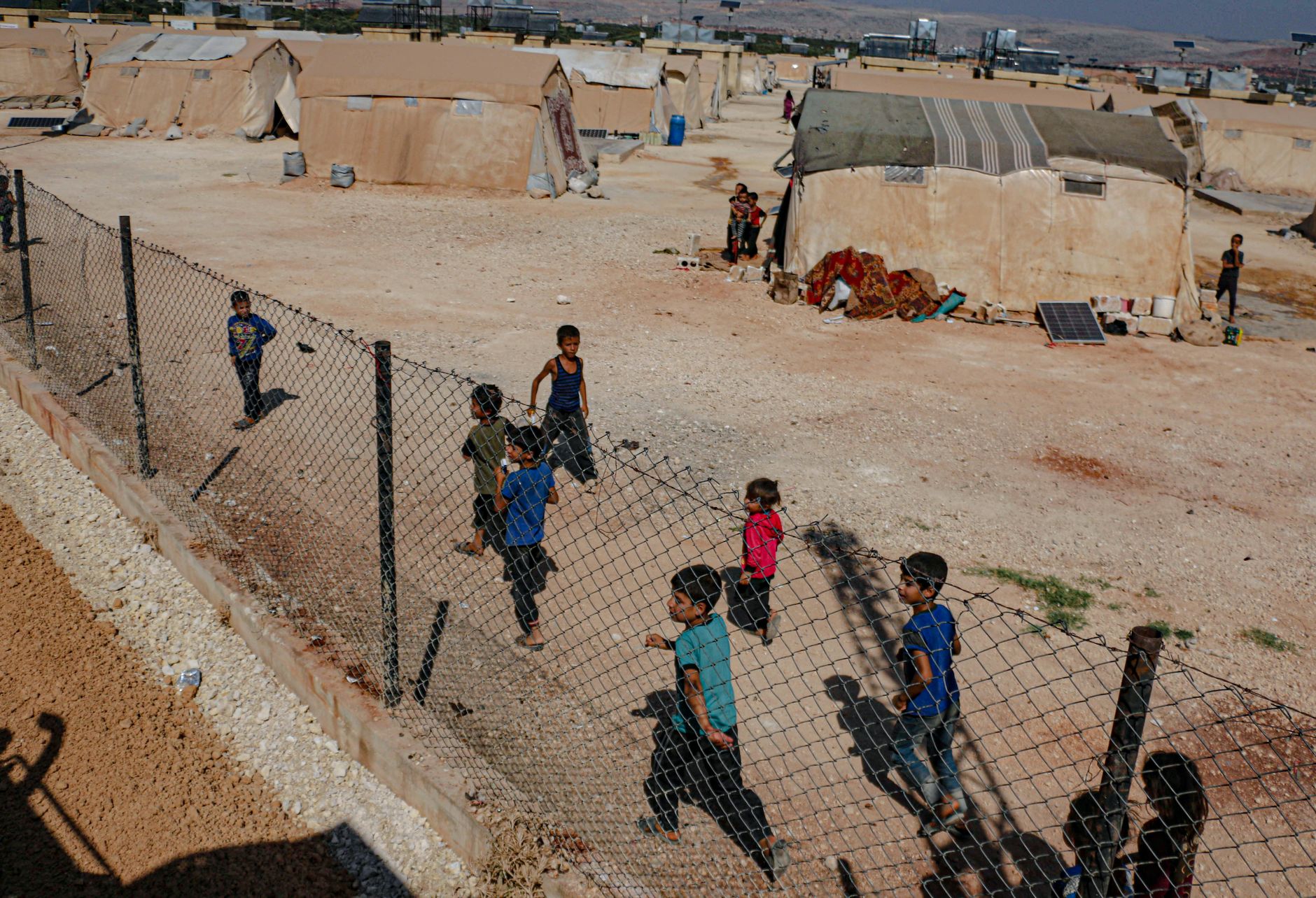 Children run along a fence in a refugee camp in Idlib, Syria, amidst tents and rugged terrain.