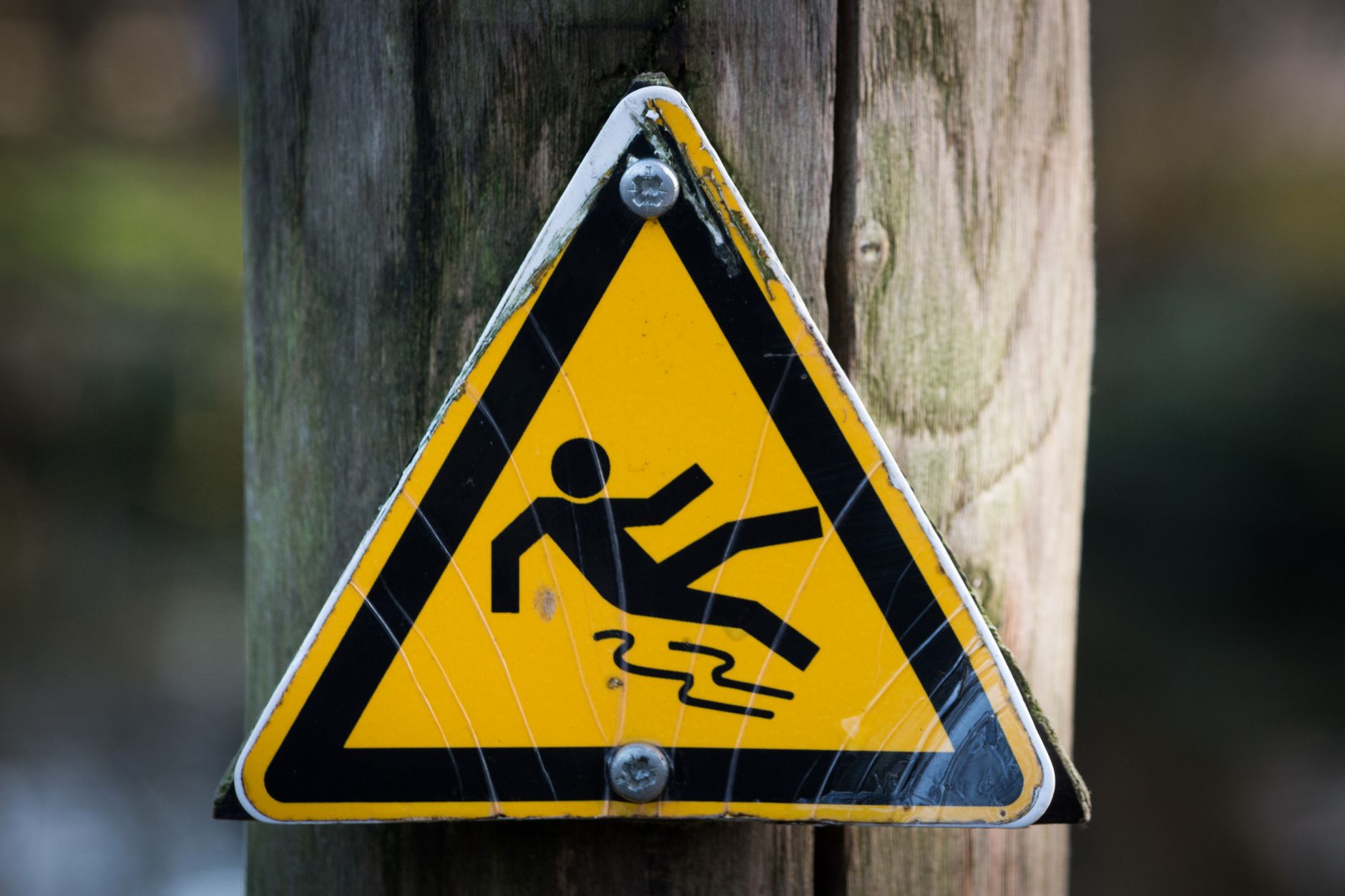 Close-up of a triangular warning sign indicating a slippery surface, fixed to a wooden post.