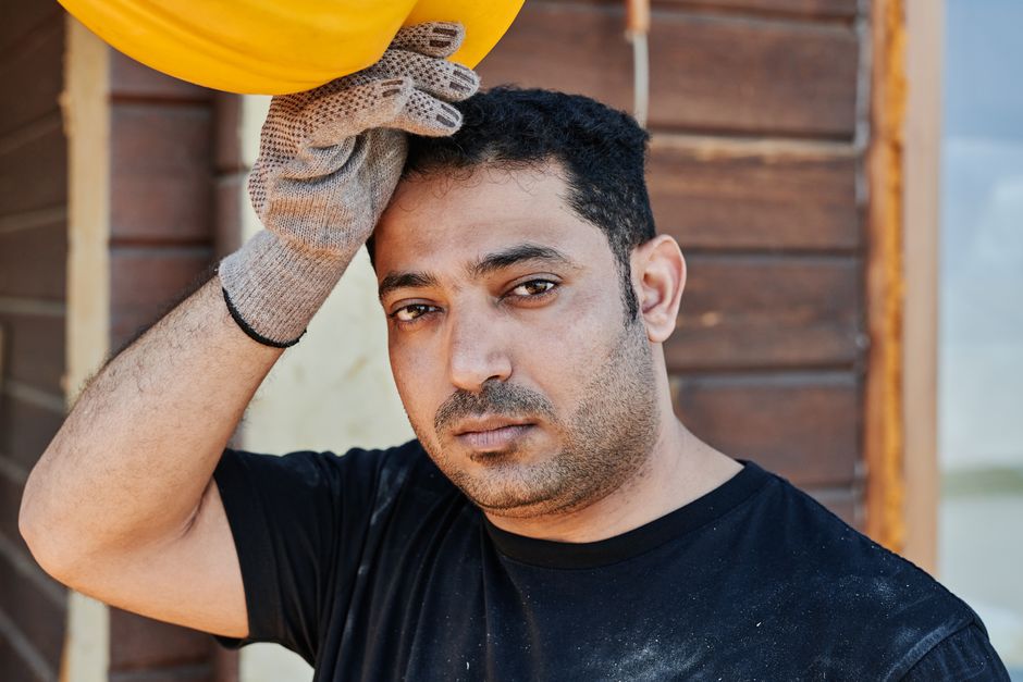 Close-up portrait of a construction worker resting, holding his hard hat. Gloved hand, thoughtful expression.