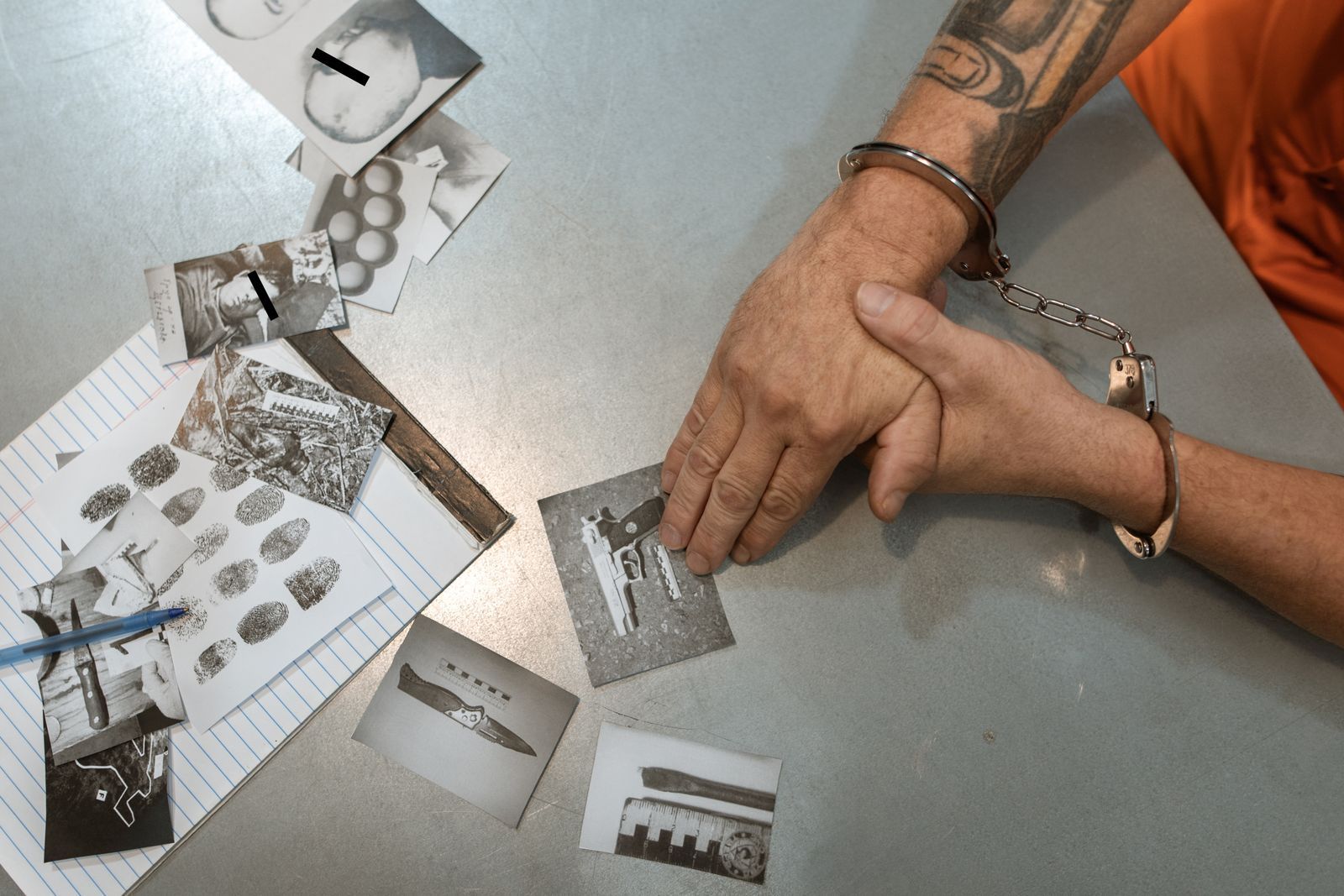 Handcuffed suspect's hands on a table beside crime scene photos and fingerprint cards