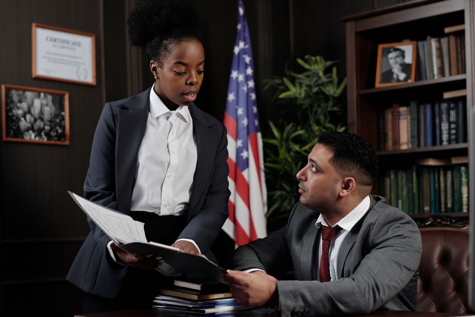 Two lawyers in suits reviewing documents at a desk in a wood-panelled law office with bookshelves