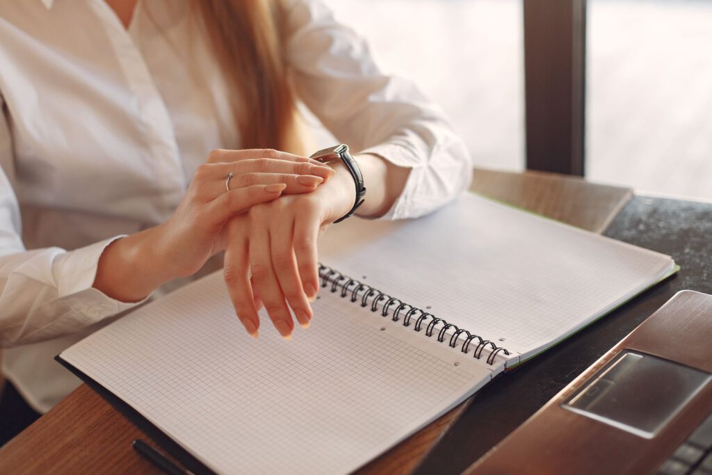 Professional in a white shirt checking her wristwatch over an open notebook and laptop at a desk