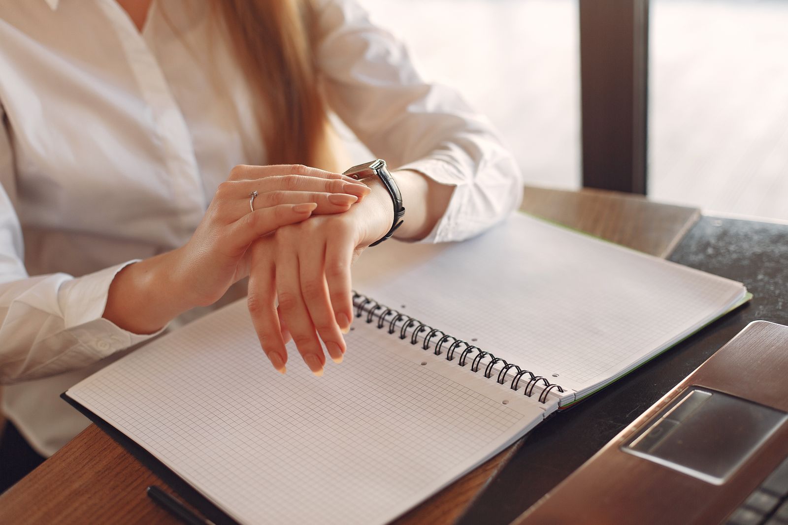 Professional in a white shirt checking her wristwatch over an open notebook and laptop at a desk