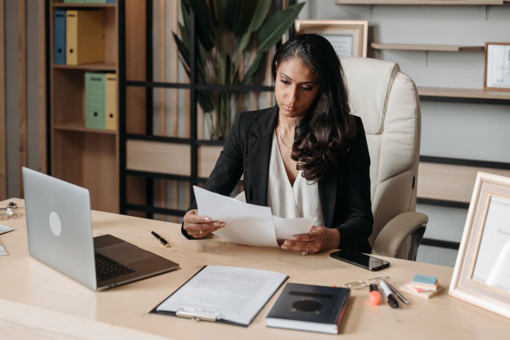 Lawyer in a black blazer reading printed documents at an office desk beside an open laptop and a clipboard