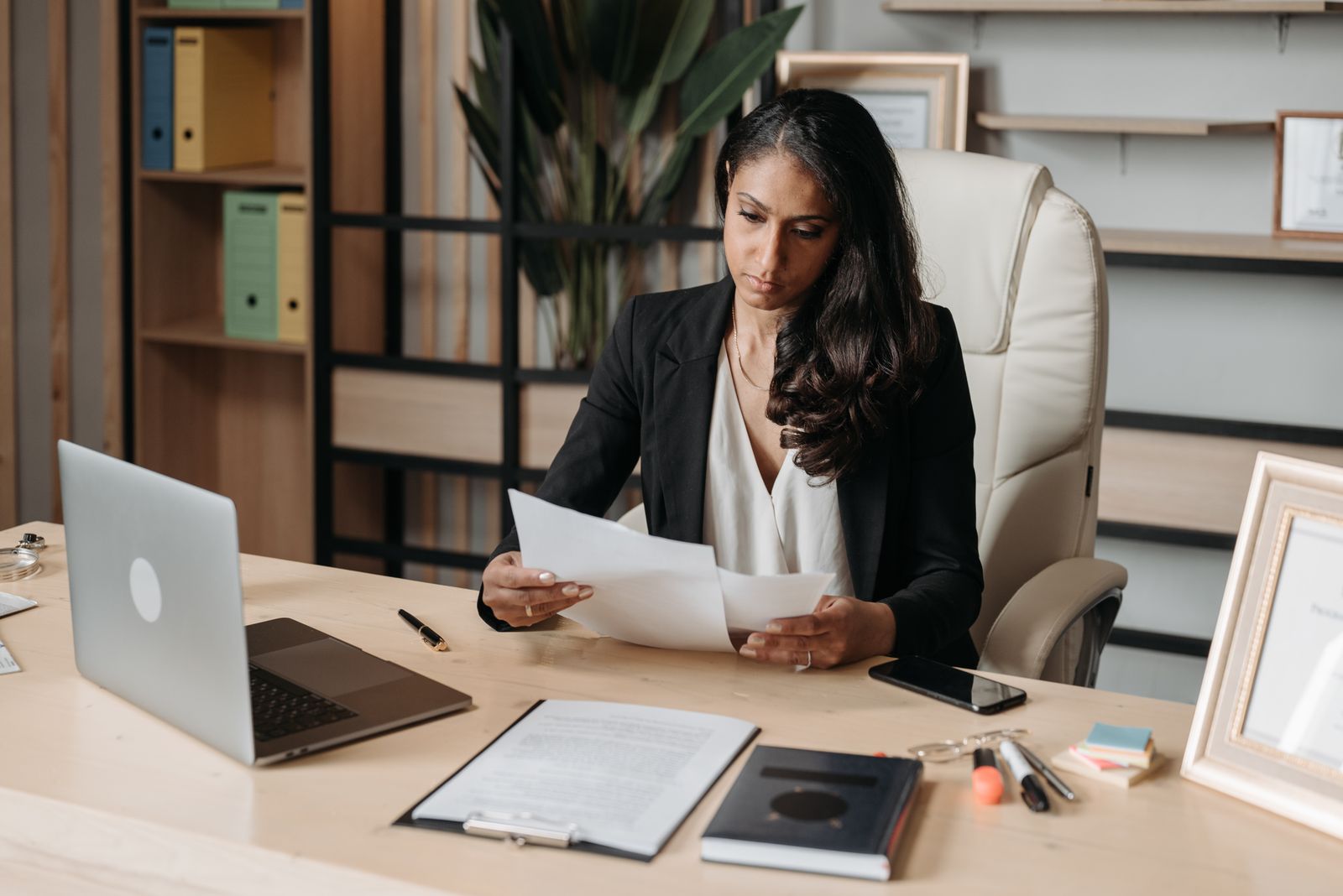 Lawyer in a black blazer reading printed documents at an office desk beside an open laptop and a clipboard