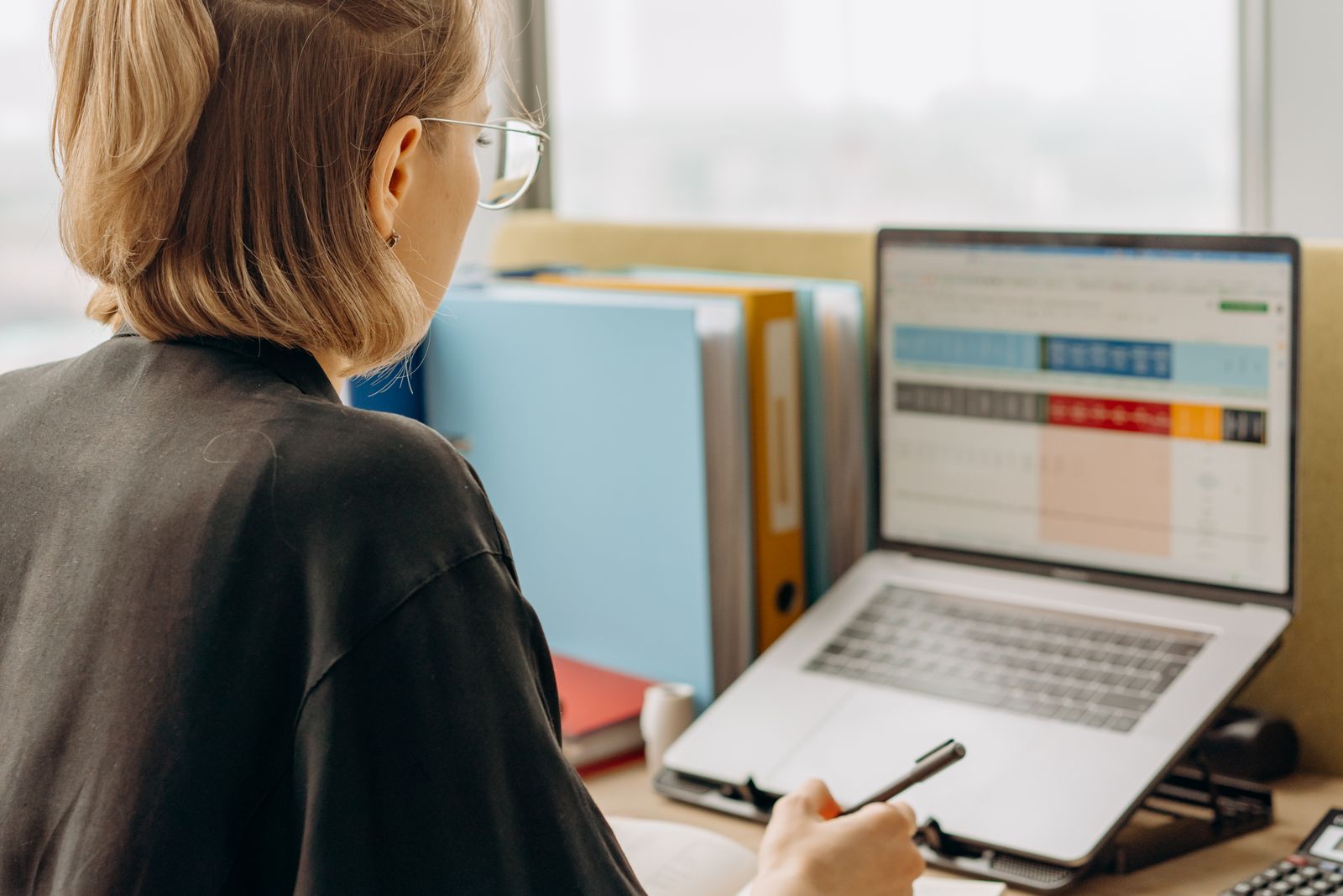Woman with glasses reviewing a colour-coded table on a laptop screen beside a row of office binders