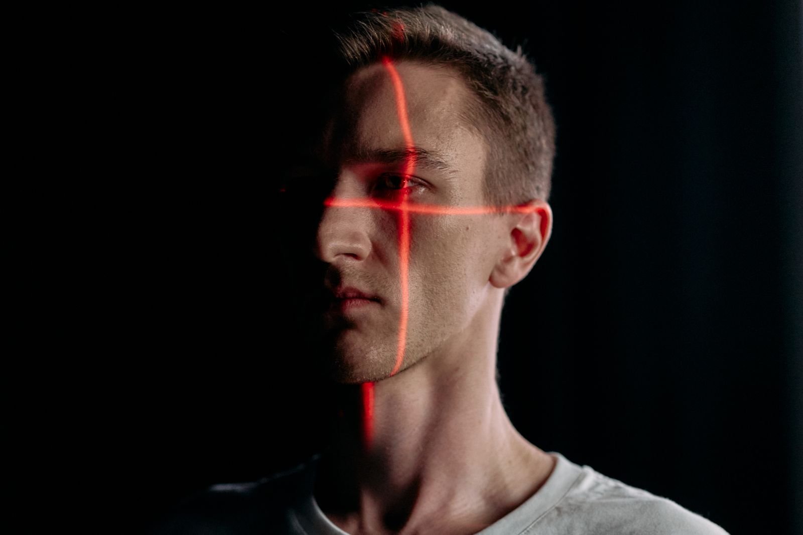 Man's face crossed by red laser lines in a dark room, suggesting biometric scanning by a machine