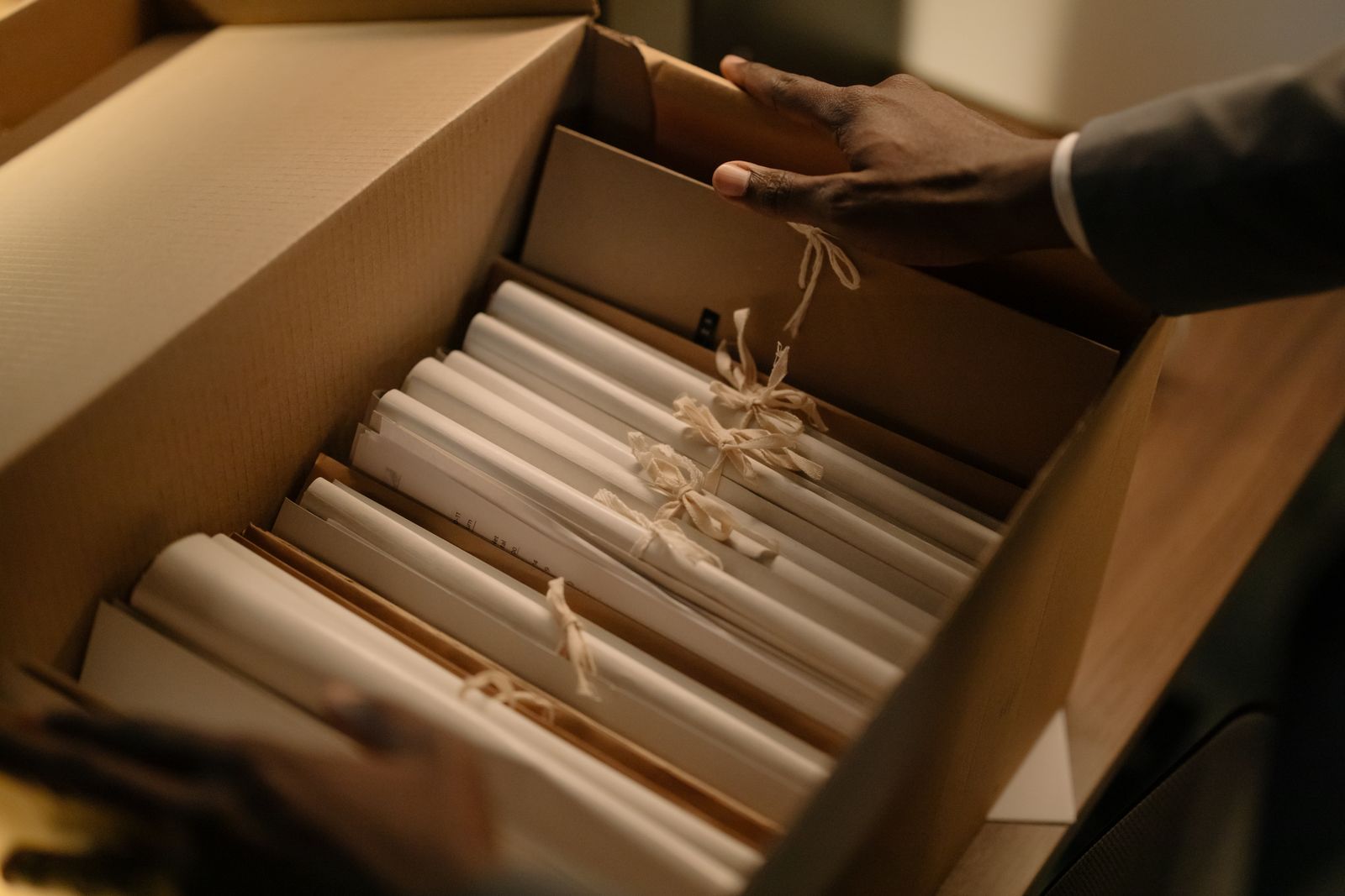 Hands sorting tied bundles of files in a cardboard archive box, like records handed over under a subpoena