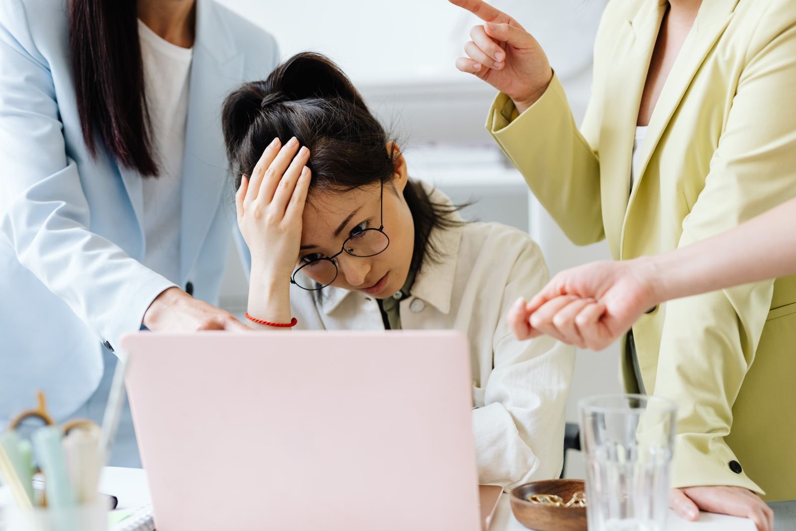 Upset office worker holding her head while colleagues point at her laptop, showing workplace harassment