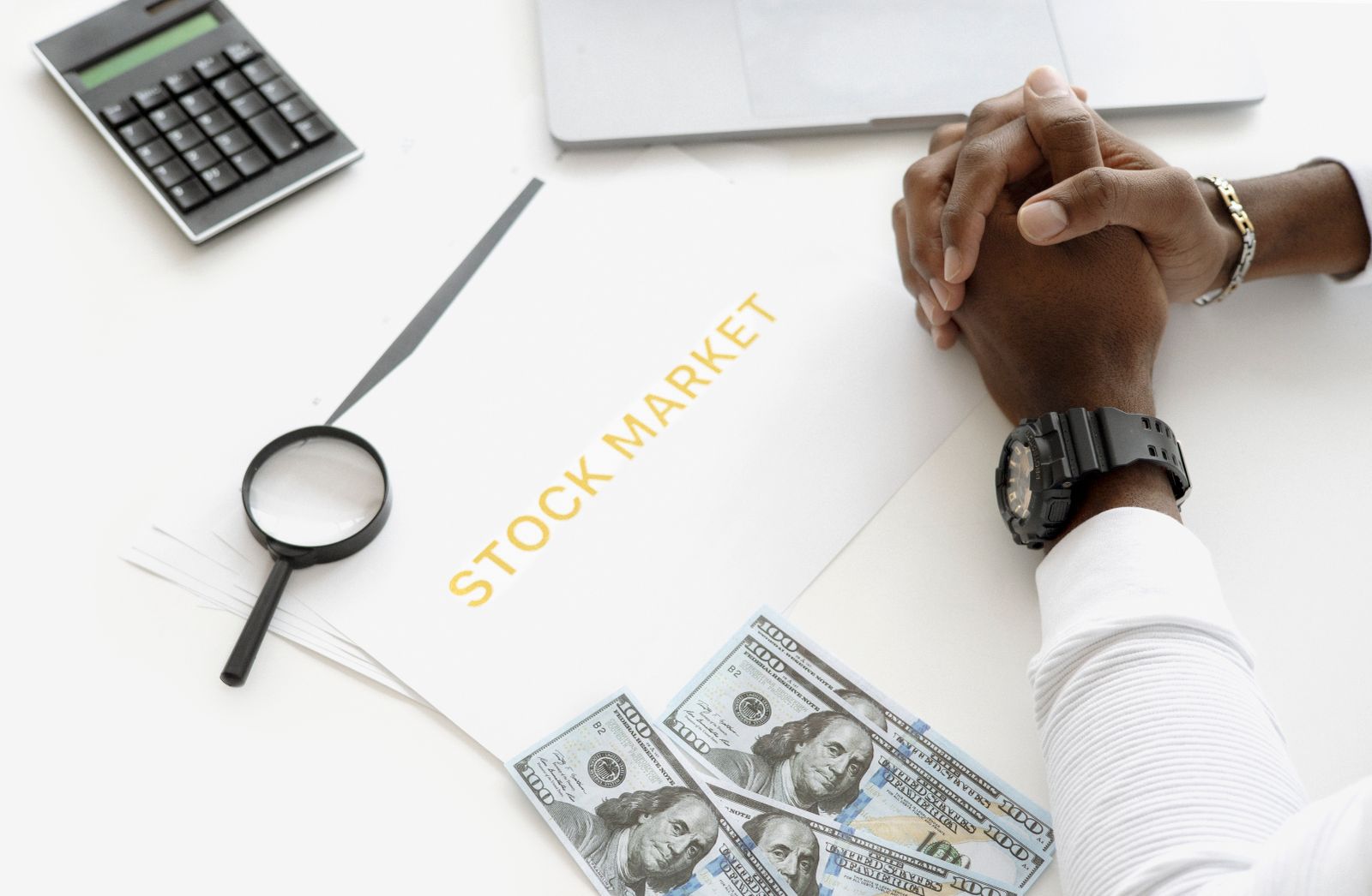 Hands folded over a stock market folder on a white desk beside a calculator, magnifying glass and dollar bills