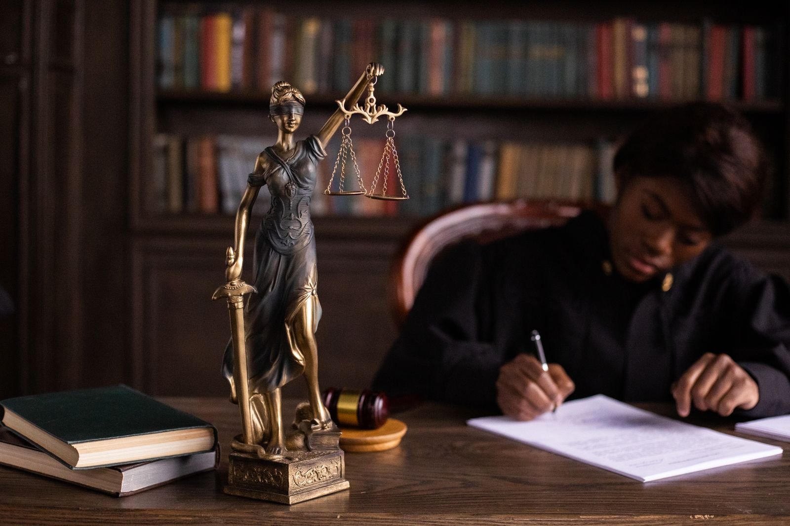 Judge in a black robe writing on documents at a desk beside a Lady Justice statue and law books