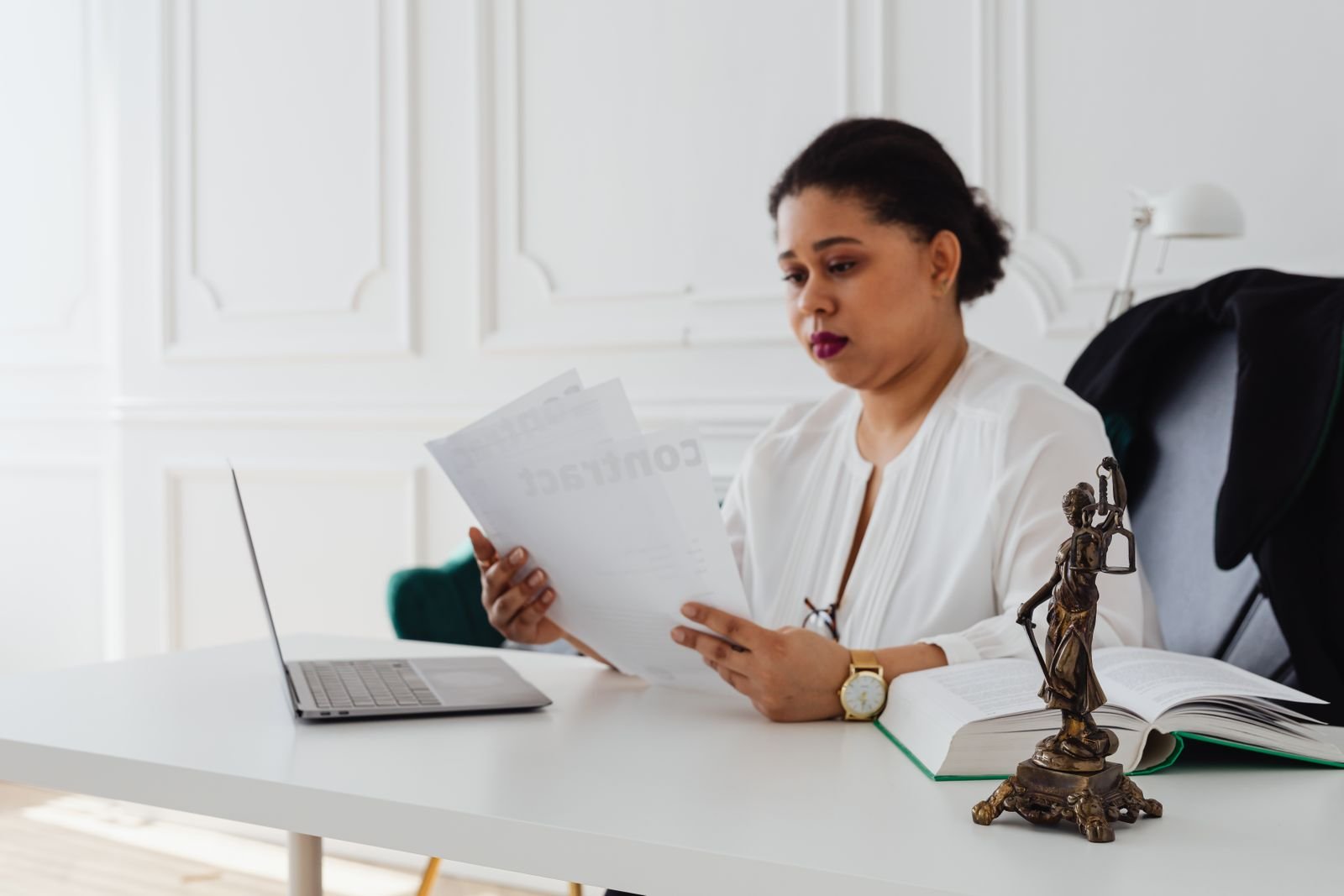Lawyer reading printed documents at a white desk with a laptop, an open law book and a Lady Justice figurine