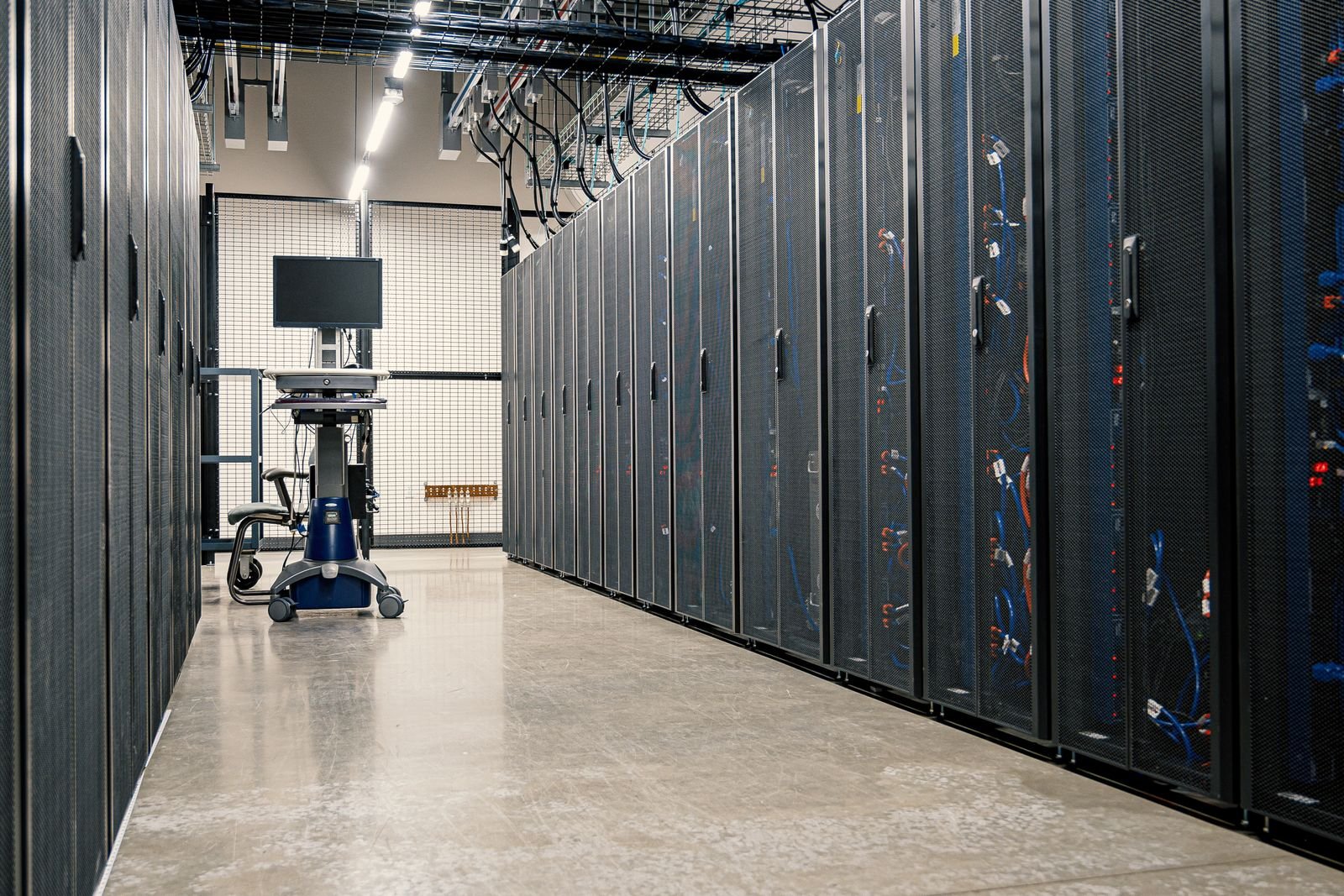 Rows of black server racks with cabling in a data centre aisle beside a computer cart