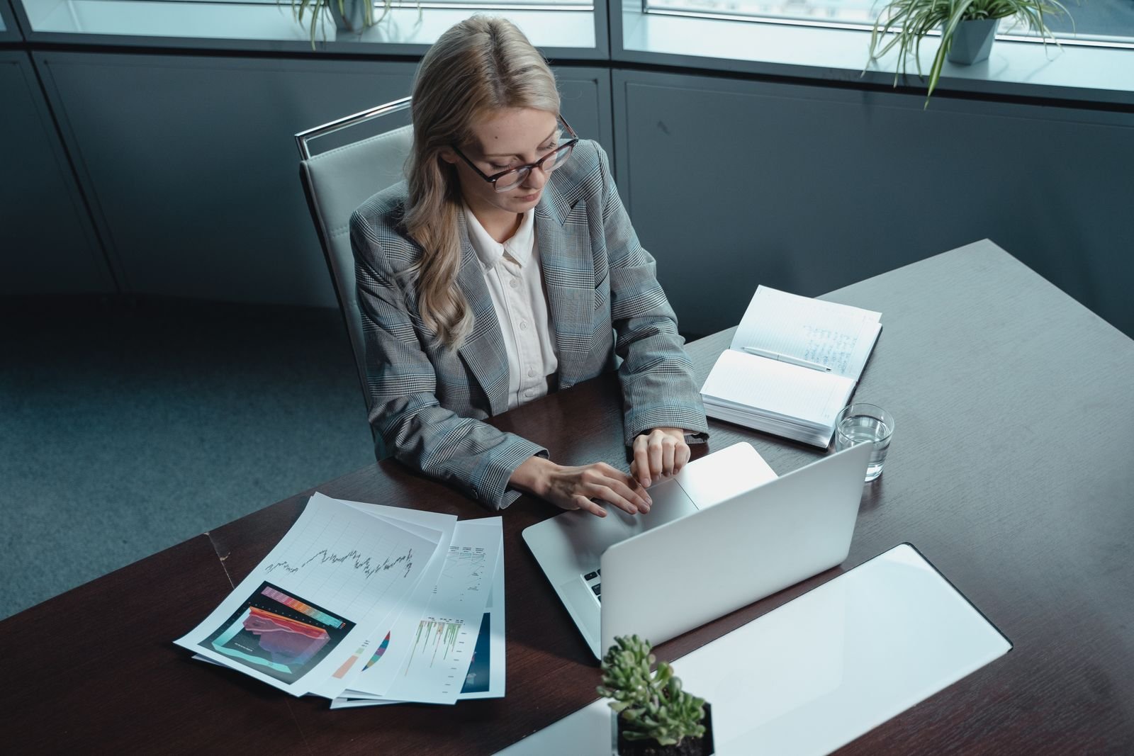 Lawyer in a grey blazer working on a laptop at an office desk beside printed reports and a notebook