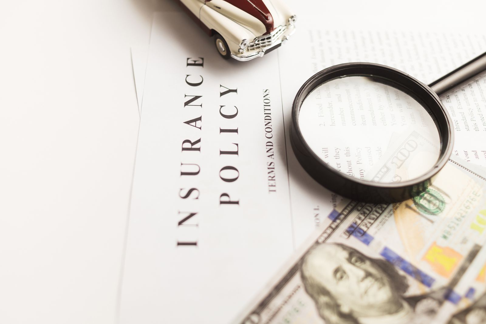 Insurance policy terms with a magnifying glass, dollar bills and a toy car on a white desk