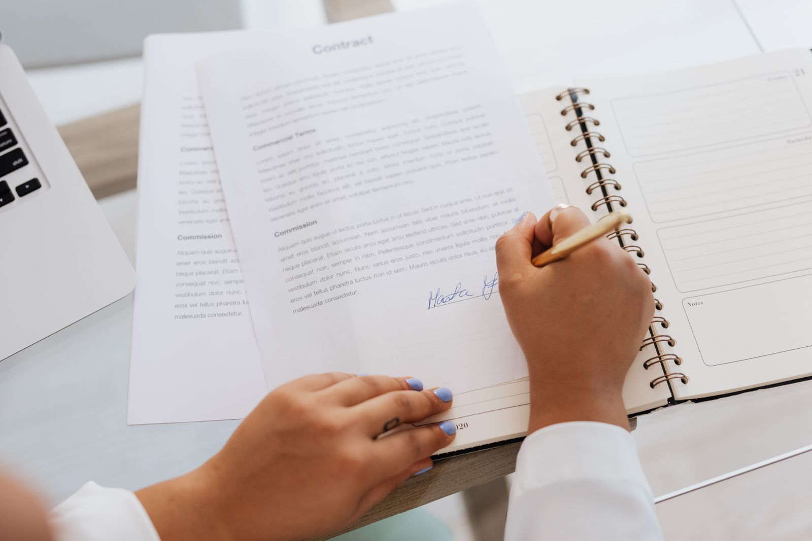 Woman signing a printed contract at a desk beside a spiral notebook and laptop