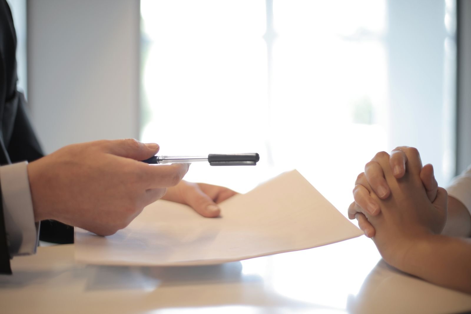 Person in a suit handing a document and pen across a desk to another person with clasped hands