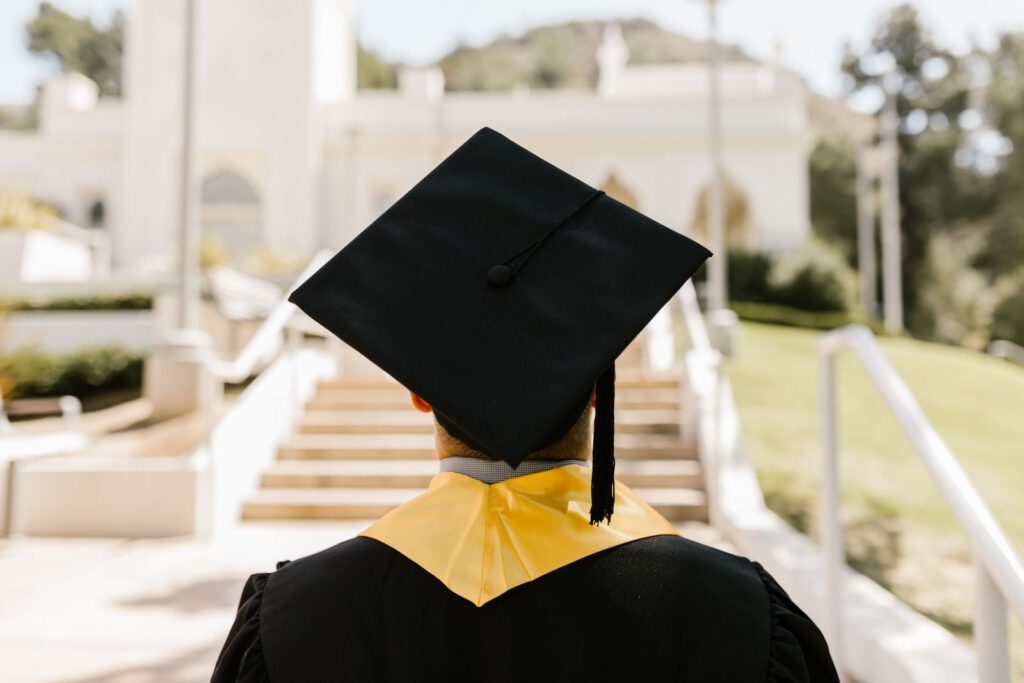 Back view of a graduate in a black mortarboard and gown with a gold stole facing campus steps