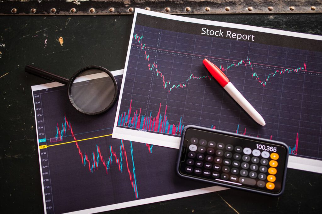 Printed stock report charts on a dark desk with a magnifying glass, red marker and a phone calculator