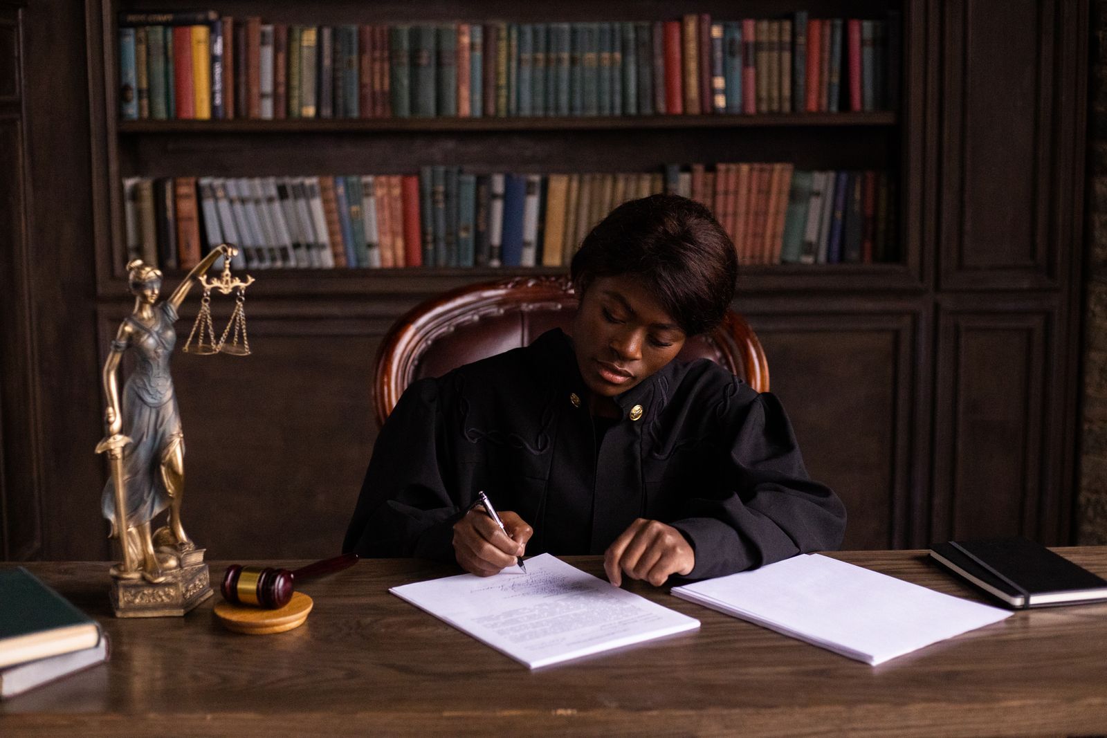 Judge in robes writing an order at a desk with a gavel and Lady Justice statue in a law library